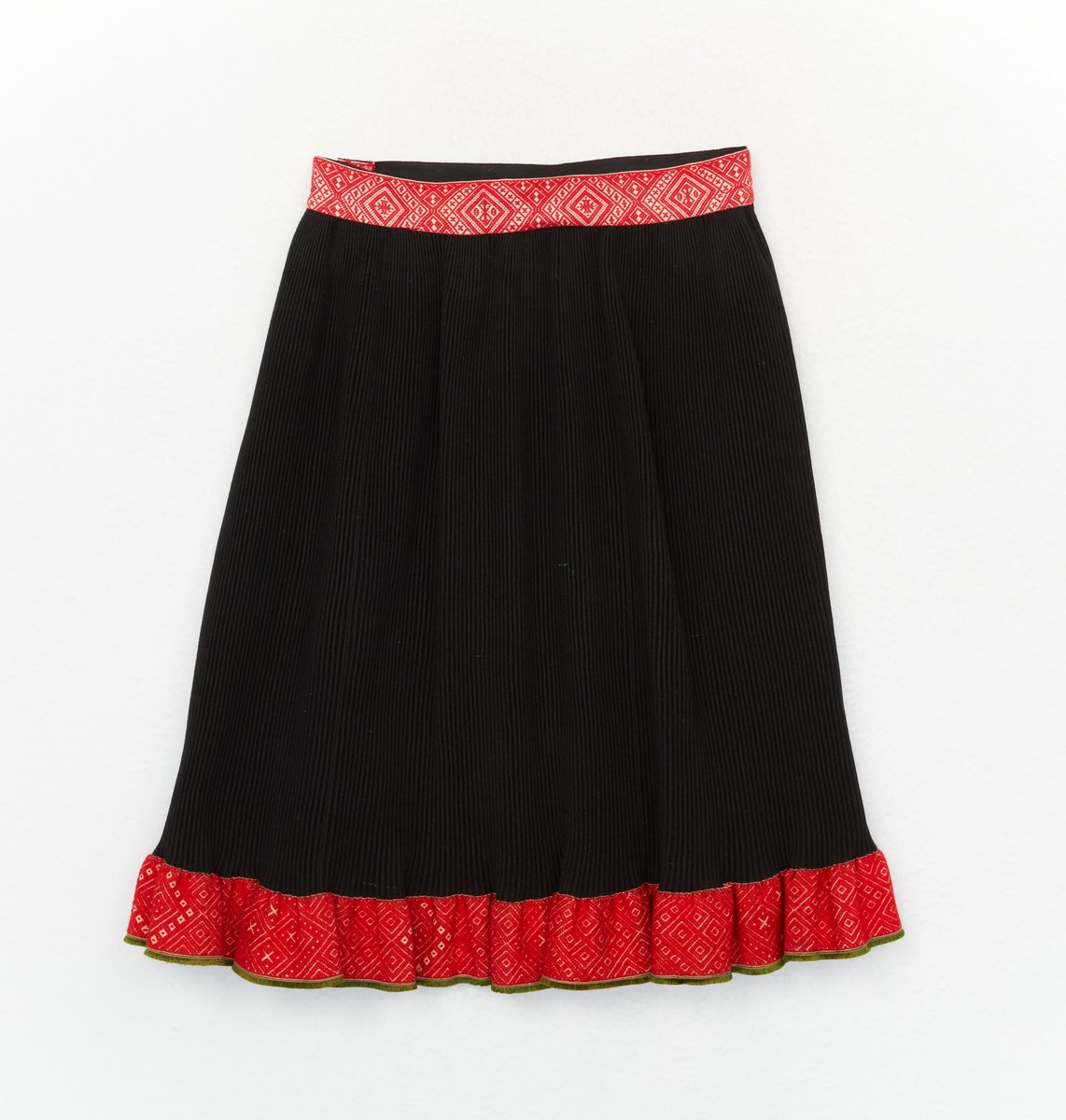 Kjol till Boda sockendräkt för kvinna. Rynkkjol av svart halvylletyg. Bredda av rött, grönt, svart och vitt tvärrandigt halvylletyg. Brett mönstervävt linningsband, knäpps i vänster sida med tre hyskor och hakar. Nedtill på rynkdelen ett brett mönstervävt band och grönt fransband.