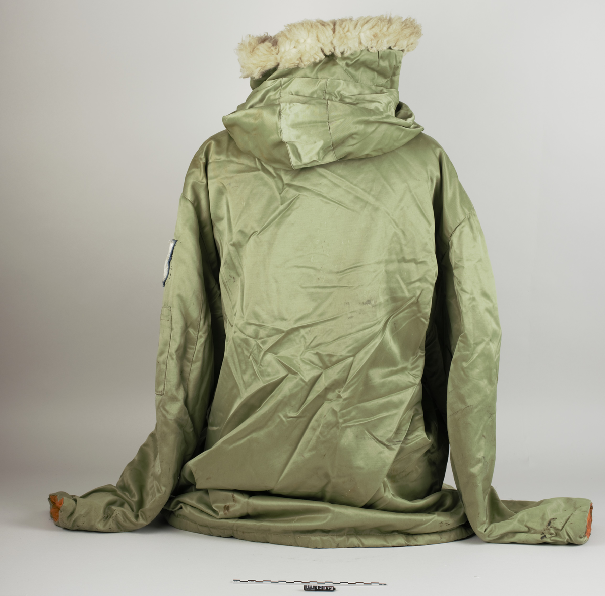 Ytterjakke i størrelse L. Jakka er vattert, av grønn syntetisk fiber, trolig nylon ytterst, og med orange bomullsfor innvendig. Foran har jakka glidelås og fire svarte knapper med tilhørende knapphull for åpning og lukking. Foran finnes fire lommer. Hver lomme festes med trykknapp. De to øverste lommene (på brystet) har påsydd svart trekant oppe og nede ved åpningen. En lomme med glidelås øverst på venstre arm. Jakka har innsydd en svart snor rundt midjen, og hette som er kantet foran med påsydd hvitgult skinn og har hempe på baksiden. Gjennom hetta går en lyseblå snor for stramming. Hetta kan ikke tas av, men er sammensydd med jakka ved et svart tøybånd. Et emblem er påsydd venstre arm. Det svært slitte emblemet har framside i blå og hvit nylon, og bakside i blå filt. Innvendig i jakkas nakke er en hempe for oppheng sydd fast, og en liten lapp med bokstaven L i rødt på hvit bakgrunn. Rett nedenfor er en større merkelapp påsydd; grå med hvit skrift, og trolig av nylon. Mesteparten av lappen har løsnet. På lappen er det trykt "Carleton manufacturers Ltèe Canada", og flere ord som er uleselige. Oljeflekker noen steder.