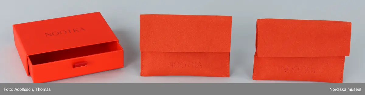 Förpackning för örhängen.
a-b) Ask av orange papp, rektangulär, med låda inskjuten från ena långsidan. Asken med handtag av tygband. Text på asken "NOOTKA".
c-d) Två påsar av orange filtat syntetmaterial. Rektangulära med flik vikt över öppningen. Text på framsidan "NOOTKA".