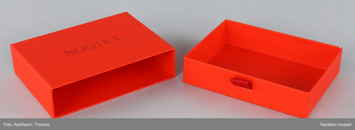 Förpackning för örhängen.
a-b) Ask av orange papp, rektangulär, med låda inskjuten från ena långsidan. Asken med handtag av tygband. Text på asken "NOOTKA".
c-d) Två påsar av orange filtat syntetmaterial. Rektangulära med flik vikt över öppningen. Text på framsidan "NOOTKA".