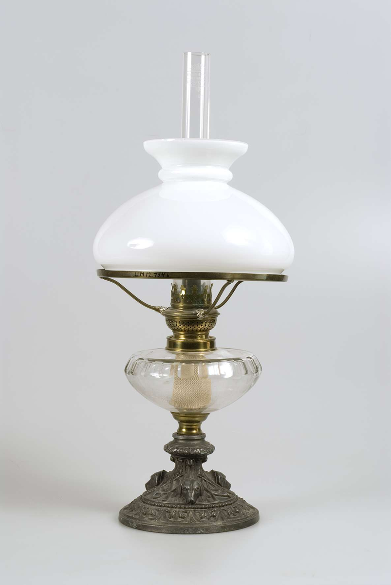 Fot av gjuten tennliknande metall med dekor i form av hundhuvuden, jaktsymboler, eklöv m.m behållare av slipat glas, brännare av mässing med text på skruven: Kosmos Brenner. Glasrör med text: Upsala Lamp och Lysoljeaffär UPSALA 10 III. Kupa av vitt mjölkglas.
Hållare för kupan söndrig. Glaskupan trasig.


 
