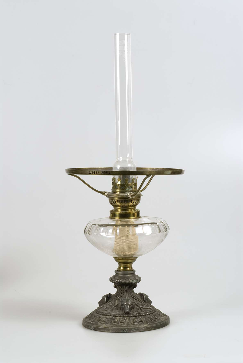 Fot av gjuten tennliknande metall med dekor i form av hundhuvuden, jaktsymboler, eklöv m.m behållare av slipat glas, brännare av mässing med text på skruven: Kosmos Brenner. Glasrör med text: Upsala Lamp och Lysoljeaffär UPSALA 10 III. Kupa av vitt mjölkglas.
Hållare för kupan söndrig. Glaskupan trasig.


 
