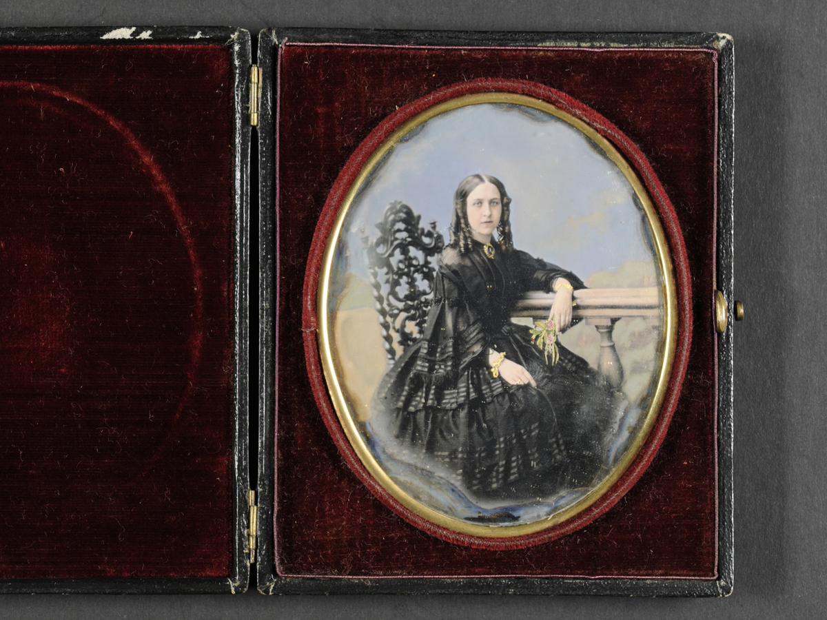 Daguerreotypi: Portrett av sittende kvinne i sort kjole (helfigur) med hengekrøller. Hun lener armen på et rekkverk med blomsterbukett i hånden.
