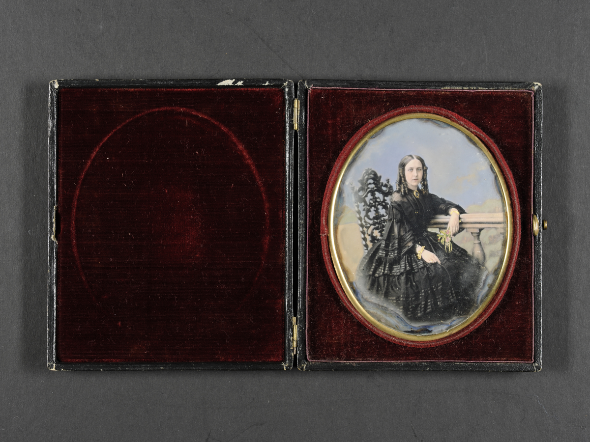 Daguerreotypi: Portrett av sittende kvinne i sort kjole (helfigur) med hengekrøller. Hun lener armen på et rekkverk med blomsterbukett i hånden.