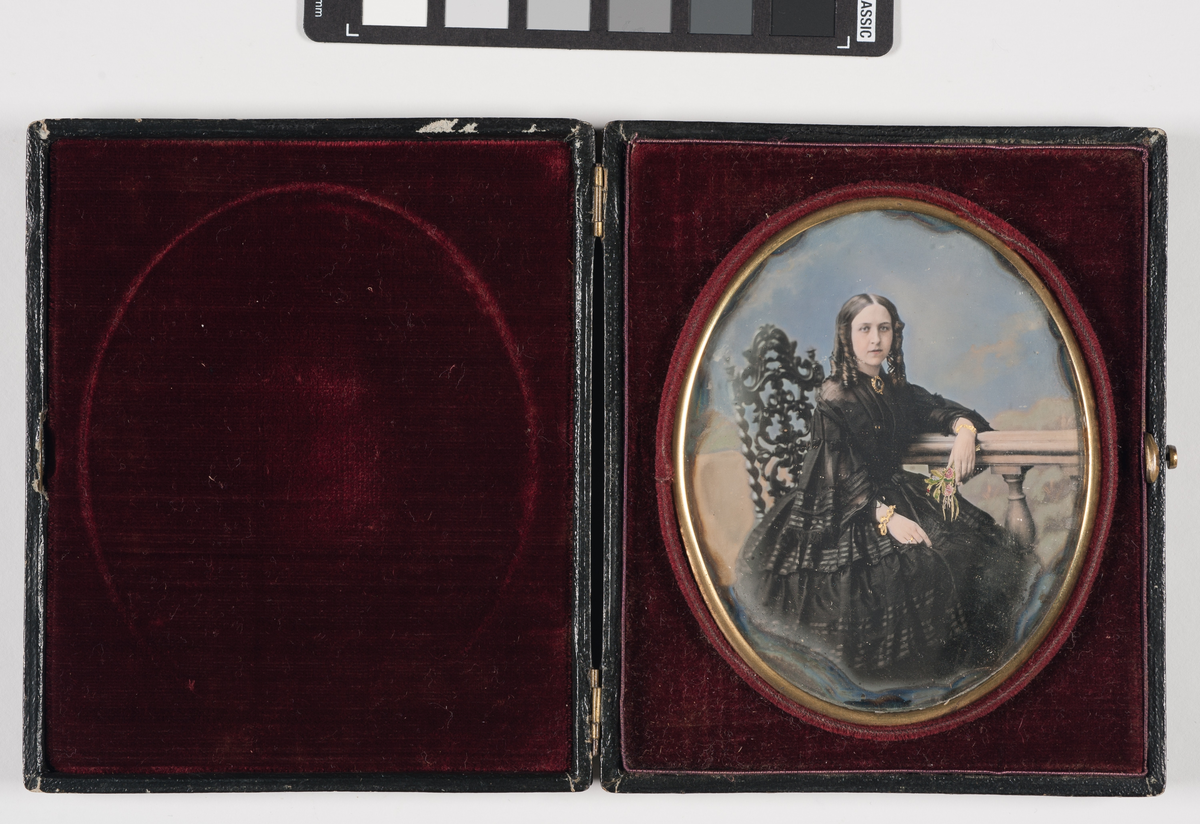 Daguerreotypi: Portrett av sittende kvinne i sort kjole (helfigur) med hengekrøller. Hun lener armen på et rekkverk med blomsterbukett i hånden.