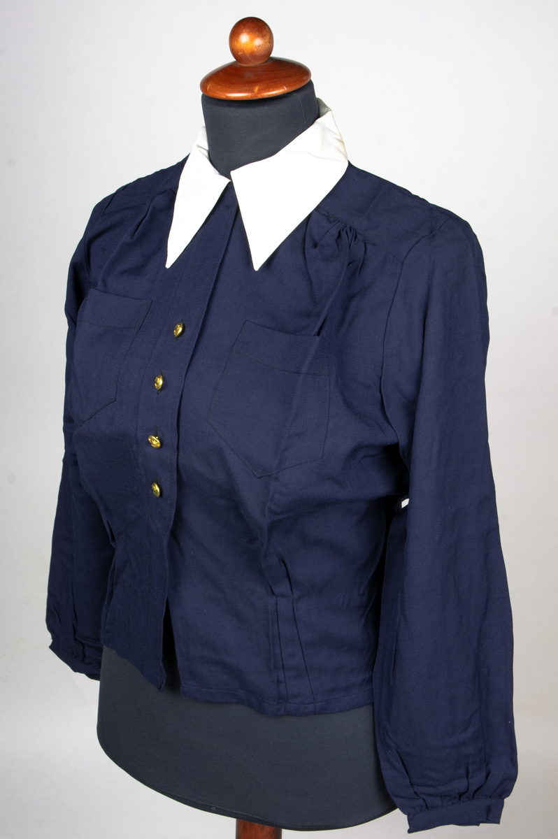 Blus av blått bomullstyg. Blusen har en vit nedvikt löskrage med osynlig knäppning. Blusen knäpps med en rad om fyra uniformsknappar. Blusen har två bröstfickor och har långa ärmar. Blusen är storlek 42. Till blusen hör en extra krage.