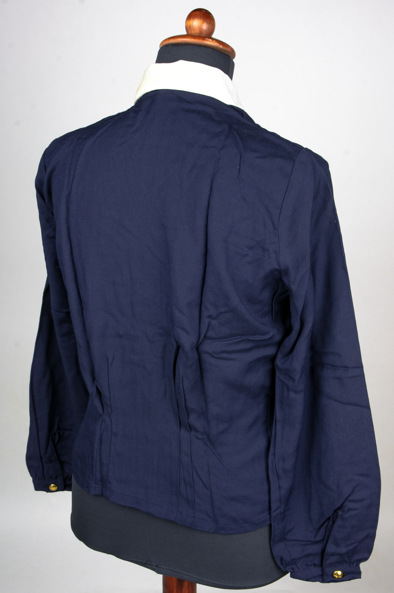 Blus av blått bomullstyg. Blusen har en vit nedvikt löskrage med osynlig knäppning. Blusen knäpps med en rad om fyra uniformsknappar. Blusen har två bröstfickor och har långa ärmar. Blusen är storlek 42. Till blusen hör en extra krage.