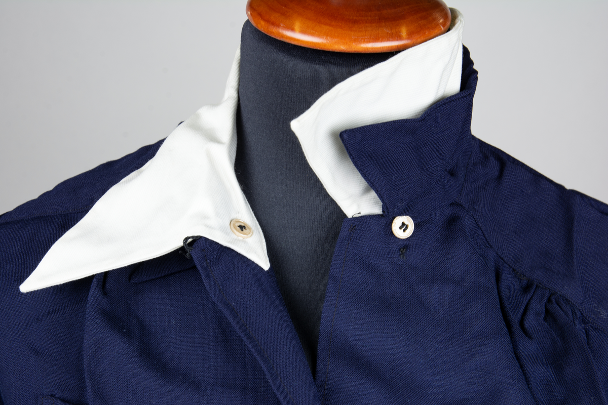 Blus av blått bomullstyg. Blusen har en vit nedvikt löskrage med osynlig knäppning. Blusen knäpps med en rad om fyra uniformsknappar. Blusen har två bröstfickor och har långa ärmar. Blusen är storlek 42. Till blusen hör en extra krage.