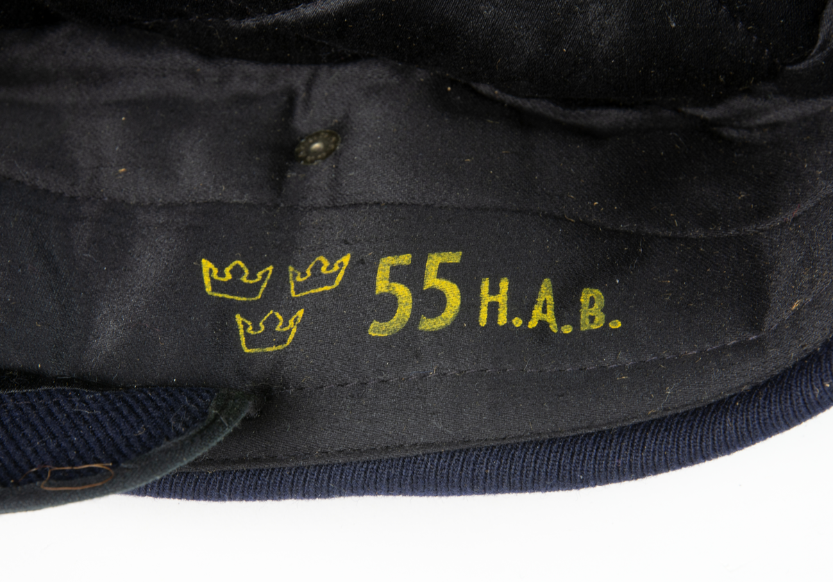 Blå vintermössa med skärm och flygemblem framtill. Baktill är ett brätte uppvikt. 
Mössan har de tre kronorna "55" och texten "H.A.B." stämplat i gult på vänster insida.
