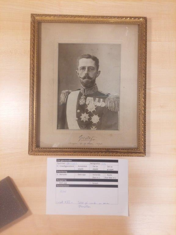 Innrammet foto med rektangulær form. Portrettet viser kronprins Gustav av Sverige. Han er fremstilt kun med overkropp, i uniform med mange ordner, med bart, hakeskjegg og lorgnett. Foto er limt på grå kartong og har gullforgylt treramme med perlebord, samt beskyttende glass. Baksiden er kledd med silkelingnende tekstil dekorert med fleur-de-lis over det hele.