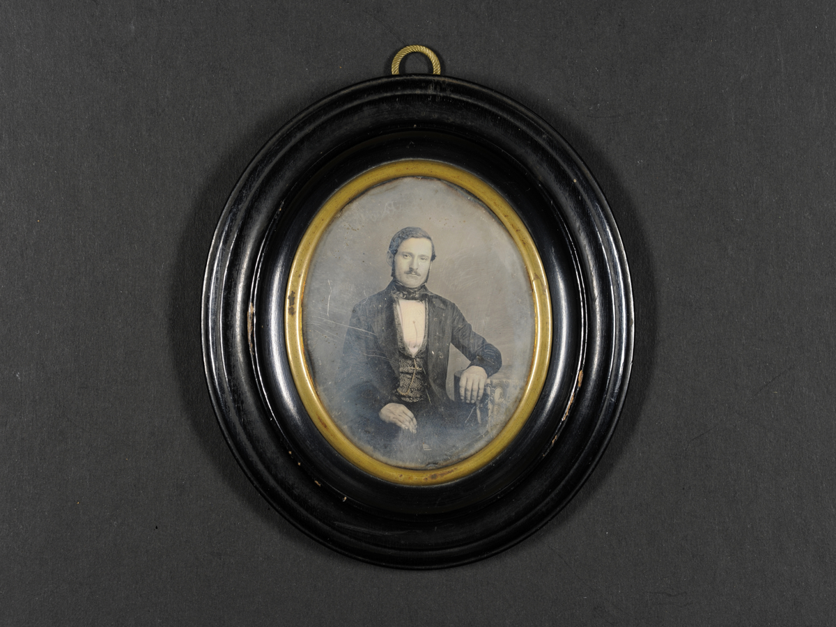 Daguerreotypi: Portrett av en mann med bart og kinnskjegg, sittende halvfigur. Mønstret vest og klokkekjede. Arm lent på et bord.