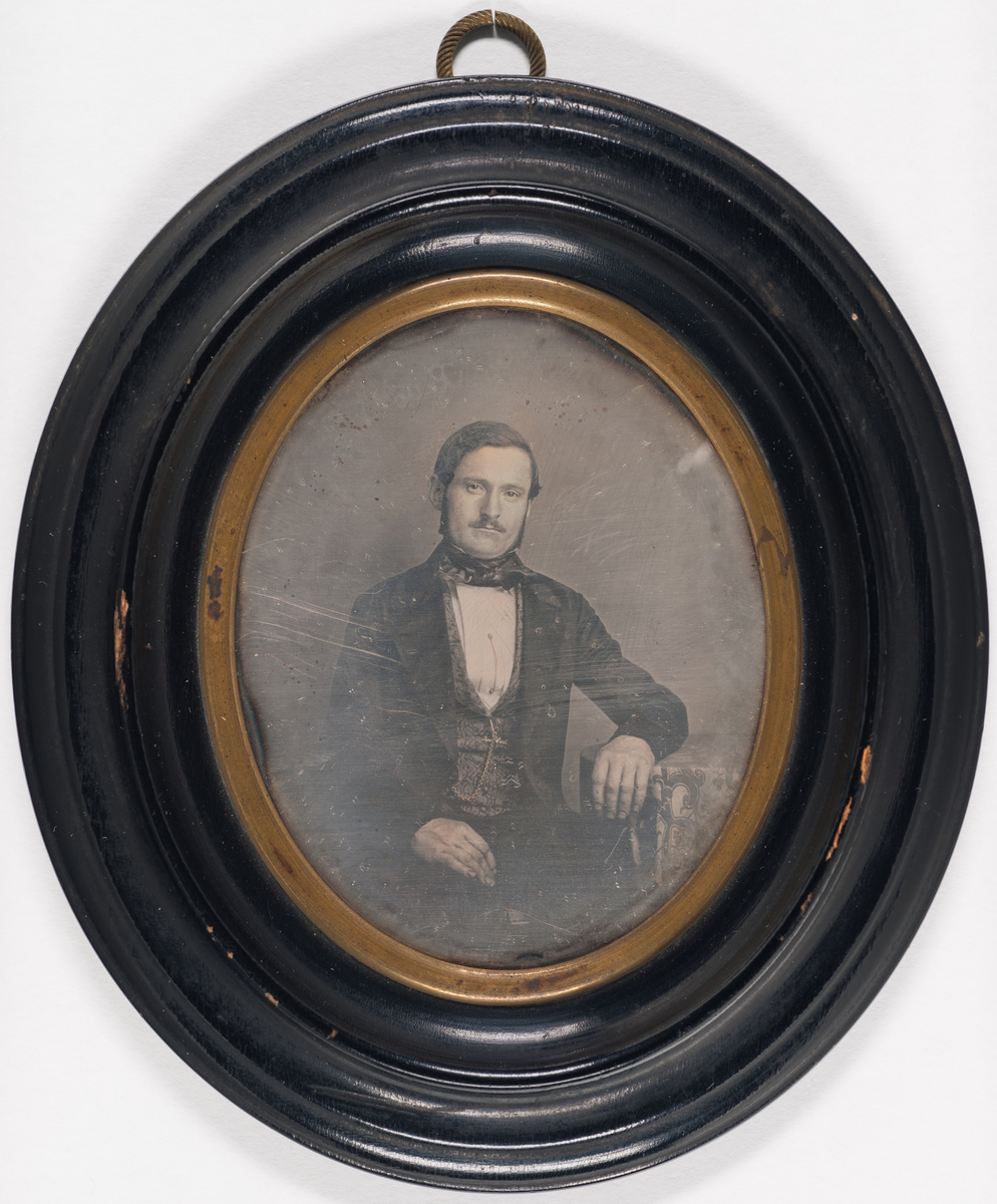 Daguerreotypi: Portrett av en mann med bart og kinnskjegg, sittende halvfigur. Mønstret vest og klokkekjede. Arm lent på et bord.