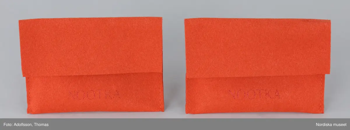 Förpackning för örhängen.
a-b) Ask av orange papp, rektangulär, med låda inskjuten från ena långsidan. Asken med handtag av tygband. Text på asken "NOOTKA".
c-d) Två påsar av orange filtat syntetmaterial. Rektangulära med flik vikt över öppningen. Text på framsidan "NOOTKA".