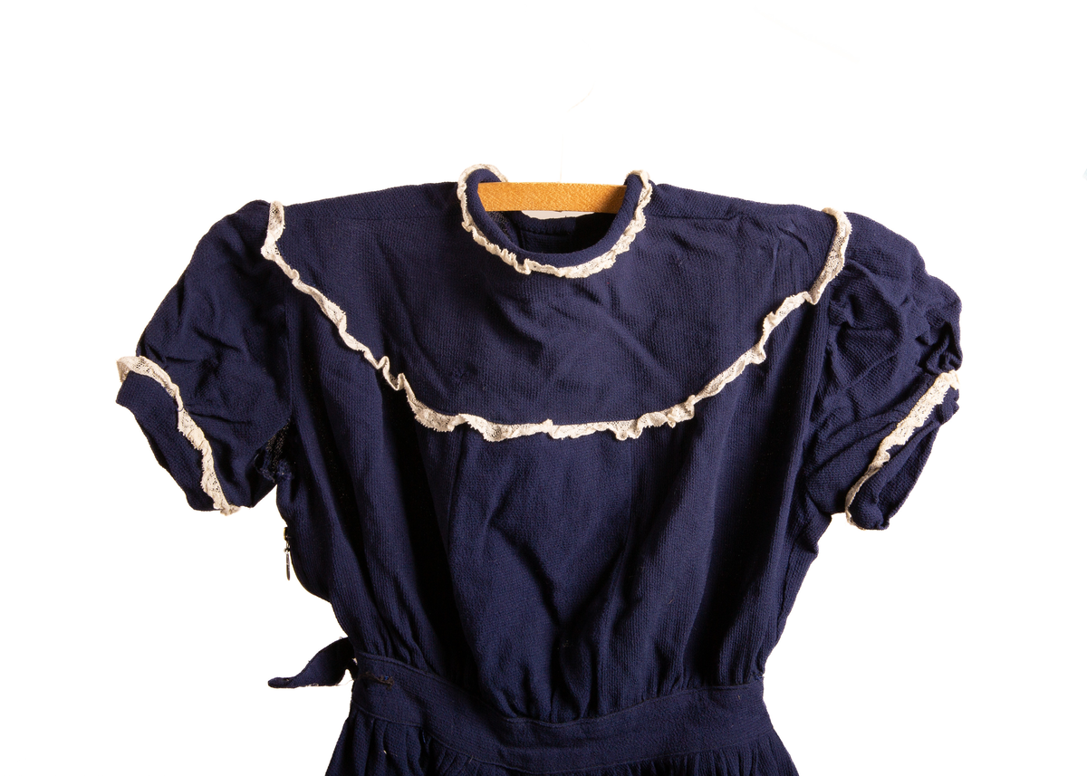 Barnekjole i blått bomullsstoff med hvite blondedekor rundt ermer og krage. Har fem tykknapper i svart metall på rygg. Glidelås på høyreside under erme og ned til midten av skjørtet. Et imitert belte over midjen som på høyreside avsluttes med et tøystykke som legger seg over glidelås og som festes med knapp (knappen mangler).

Maskinsøm med blå tråd.