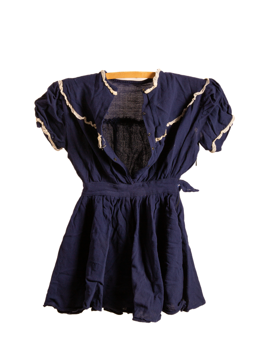 Barnekjole i blått bomullsstoff med hvite blondedekor rundt ermer og krage. Har fem tykknapper i svart metall på rygg. Glidelås på høyreside under erme og ned til midten av skjørtet. Et imitert belte over midjen som på høyreside avsluttes med et tøystykke som legger seg over glidelås og som festes med knapp (knappen mangler).

Maskinsøm med blå tråd.