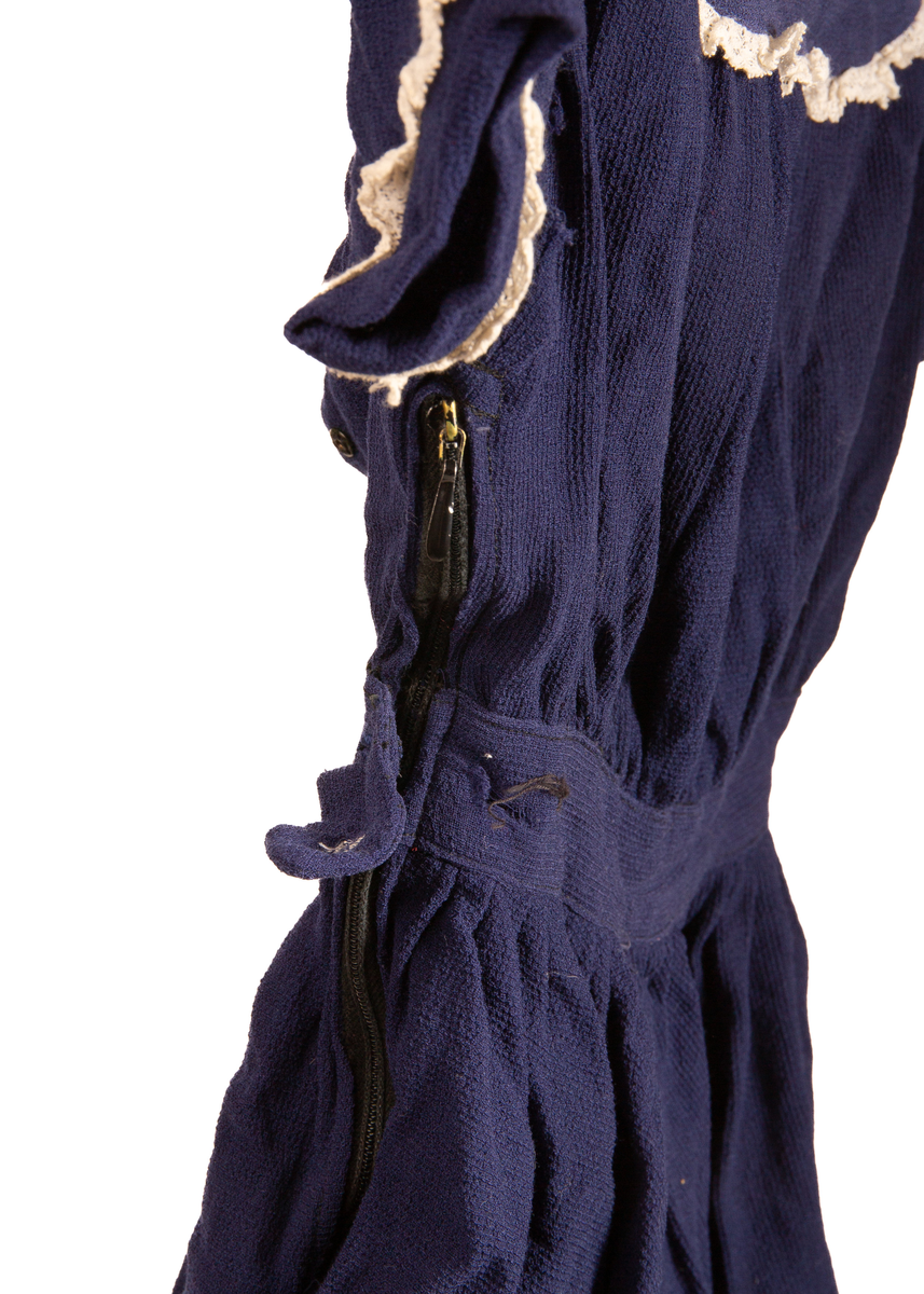 Barnekjole i blått bomullsstoff med hvite blondedekor rundt ermer og krage. Har fem tykknapper i svart metall på rygg. Glidelås på høyreside under erme og ned til midten av skjørtet. Et imitert belte over midjen som på høyreside avsluttes med et tøystykke som legger seg over glidelås og som festes med knapp (knappen mangler).

Maskinsøm med blå tråd.