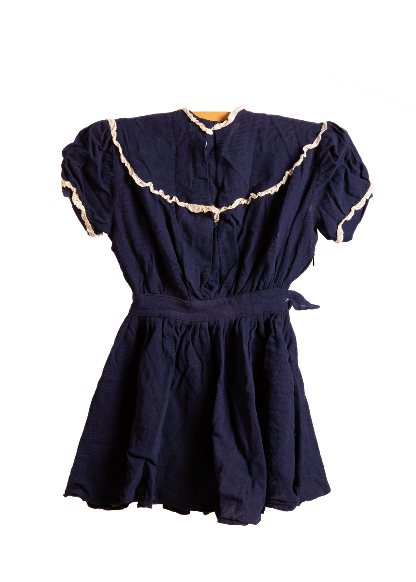 Barnekjole i blått bomullsstoff med hvite blondedekor rundt ermer og krage. Har fem tykknapper i svart metall på rygg. Glidelås på høyreside under erme og ned til midten av skjørtet. Et imitert belte over midjen som på høyreside avsluttes med et tøystykke som legger seg over glidelås og som festes med knapp (knappen mangler).

Maskinsøm med blå tråd.