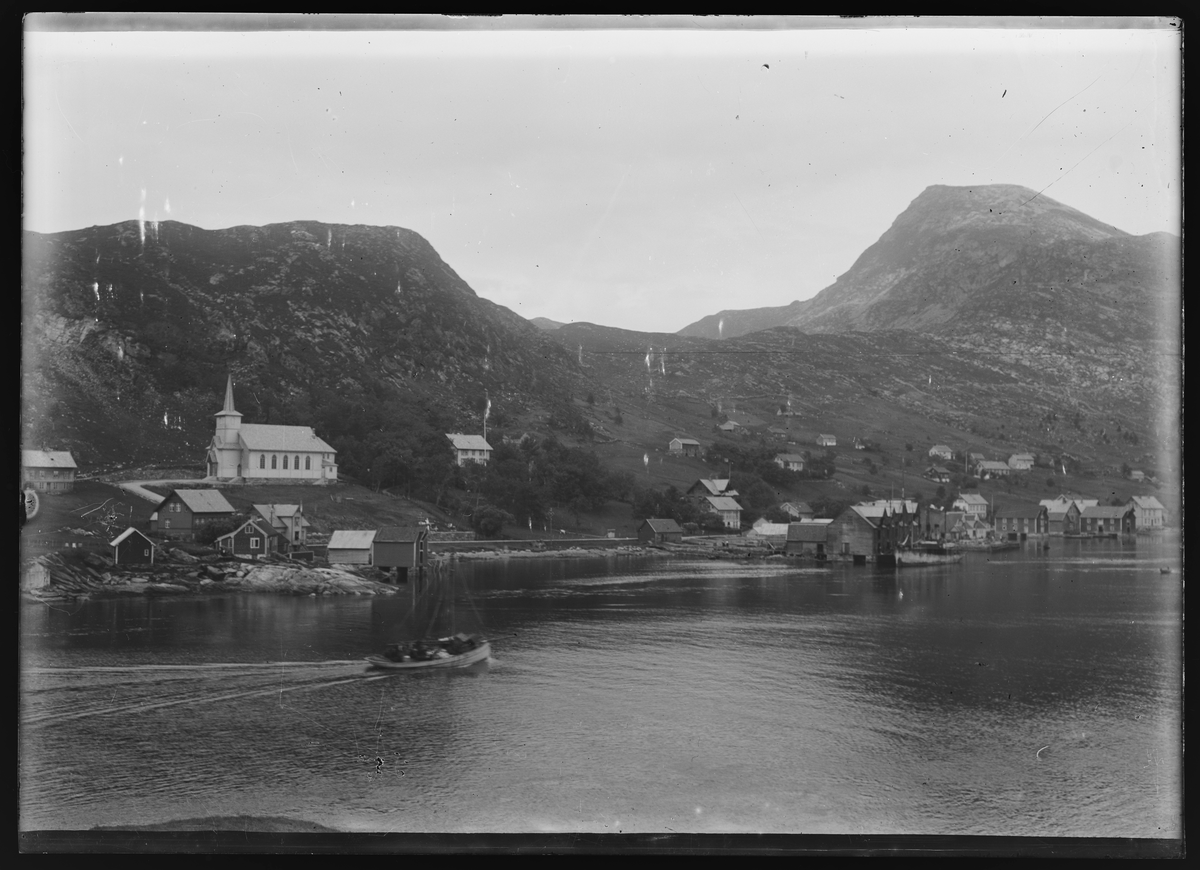 Bebyggelse nær sjøen. På vannet i forgrunnen kjører det en båt. Fra bygningene i bakgrunnen går det oppover mot noen åser.
