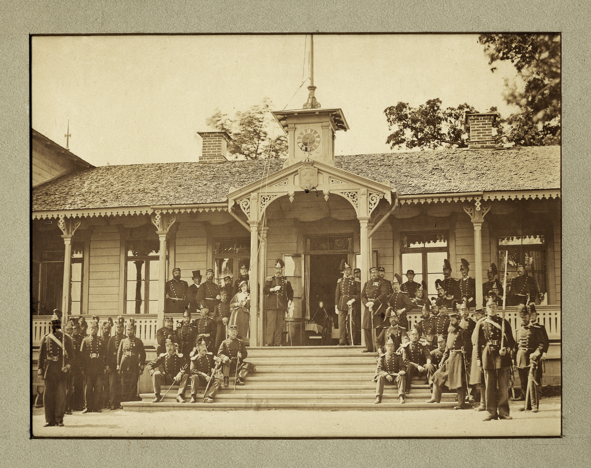 Ett antal officerare och en marketenterska har samlats utanför en officerspaviljong.
"Invigningen av Kungl. Lif Regementets Grenadier Corps' officerspaviljong å Utnäs löt, midsommardagen 1872.
Fr.v.: distinktionskorpraler, underofficerare: Övre raden: löjtnant C.A.M. Posse, reg.pastor S.J. Karlsson, kapten M. Norlin, auditör C.E. Stålhös, kapten S. Creutz. Undre raden: löjtnant G. H. Bennet, kapten C.F. Klingenstierna, kapten Thure Ehrencrona, löjtnant C.E. Holmin, marketenterska. I mitten sec.chefen överste H. Falkenberg, överstelöjtnant I.W. Schenström, kapten C.G. von Ehrenheim, kapten M.O. Wrangel, underlöjtnant A.M. Nordenskjöld, underlöjtnant P.A. Rönqvist, underlöjtnant J.C Lewenhaupt, underlöjtnant C.T. Tamm, reg.intendent L.G.C. Söderqvist, underlöjtnant C.W. Kantzow, underlöjtnant G.W.E. Bjuggren, underlöjtnant C.H. Adelborg, underlöjtnant C.W.M. Kihlstedt, reg.kvart.mästare löjtnant Paul Ehrencrona".