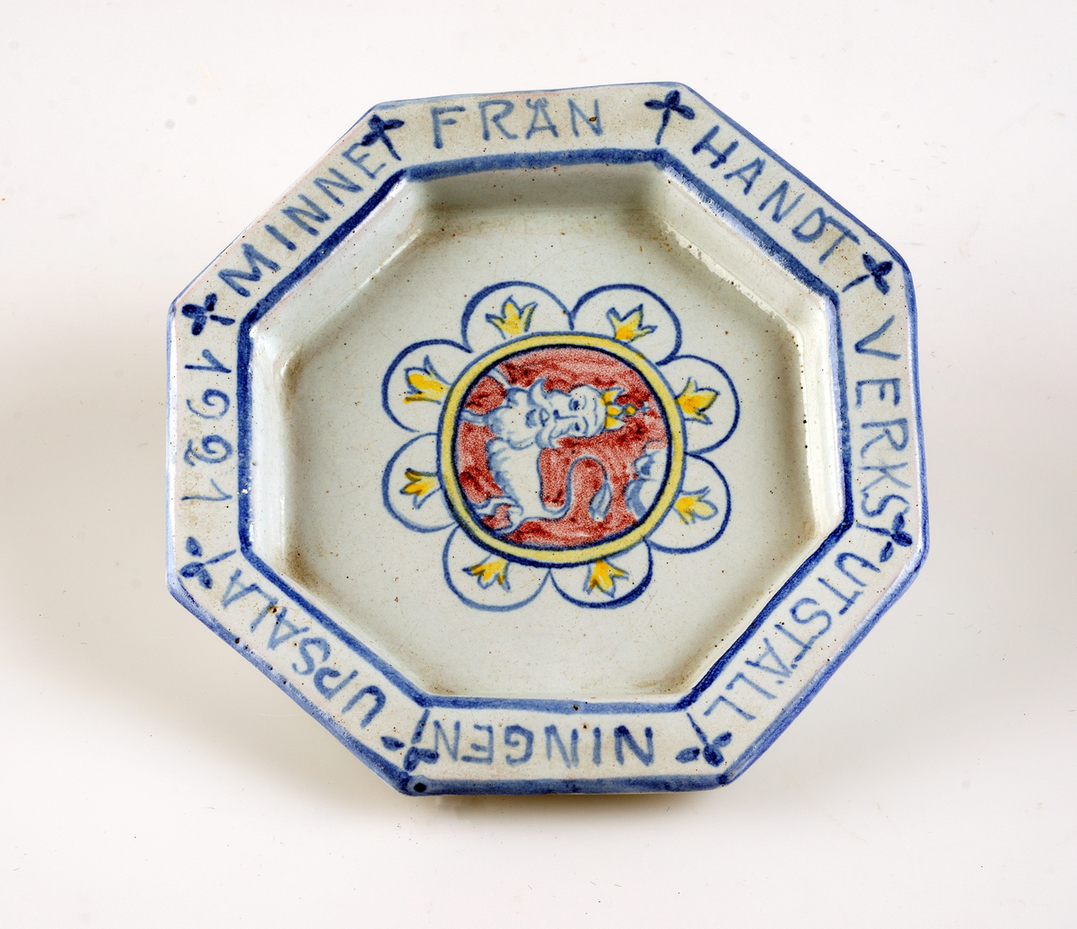 Av glaserat lergods. Dekor i blått, rött och gult på blåaktig botten. Text  "MINNE FRÅN HANTVERKSUTSTÄLLNINGEN UPSALA 1921".

