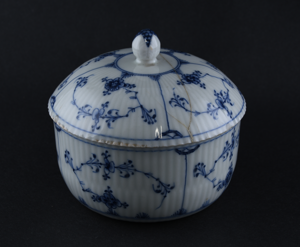 Sukkerskål av glassert porselen, med lokk. Skålen har hvit grunnfarge, og håndmalt dekor av blått stråmønster, eller musselmaling. Skålen og lokket er kannelert på utsiden. En knott på lokket har form nesten som spisskål. Lokket har knekt i to og bollen har avknekt et skår, begge er limt sammen igjen.