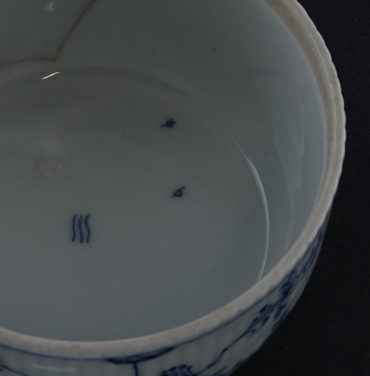 Sukkerskål av glassert porselen, med lokk. Skålen har hvit grunnfarge, og håndmalt dekor av blått stråmønster, eller musselmaling. Skålen og lokket er kannelert på utsiden. En knott på lokket har form nesten som spisskål. Lokket har knekt i to og bollen har avknekt et skår, begge er limt sammen igjen.