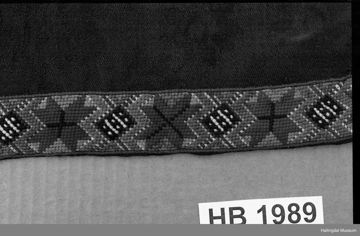 Sort ulldamask. Nederst bånd sydd på stramei. Røde åttebladroser og rutemønster i blått, lilla, orange, grønt, sort, hvitt. Øverst hektebole av blomstrete bånd.