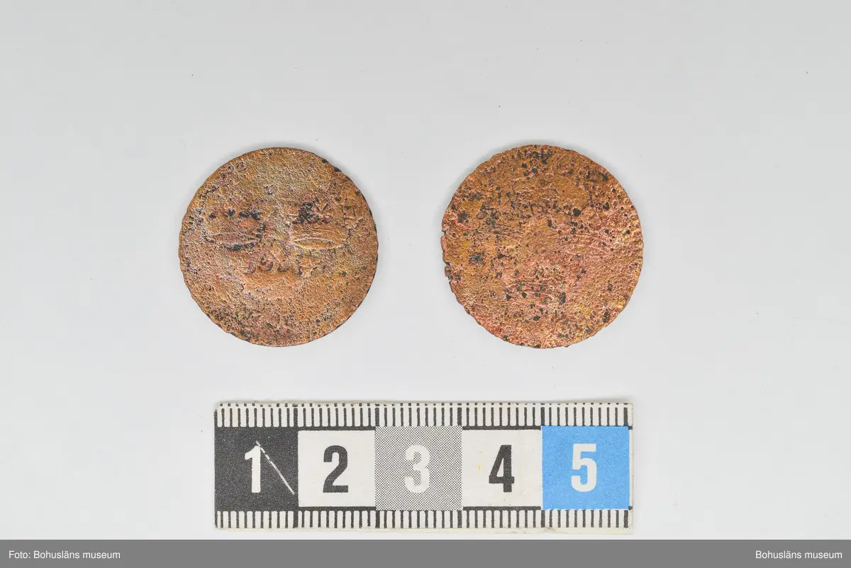 Mynt, två st. Troligen är de från år 1719 eller 1720 präglade under Ulrika Eleonoras regeringstid. De har hårt bruksslitage och hittades på en stenpackning ca 1,4 m under markytan i schakt 5, i östra delen av Kungsgatan, Uddevalla.