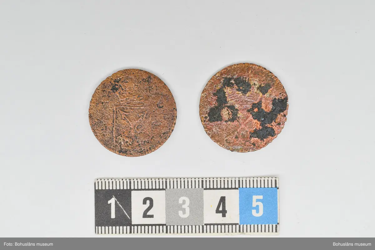Mynt, två st. Troligen är de från år 1719 eller 1720 präglade under Ulrika Eleonoras regeringstid. De har hårt bruksslitage och hittades på en stenpackning ca 1,4 m under markytan i schakt 5, i östra delen av Kungsgatan, Uddevalla.