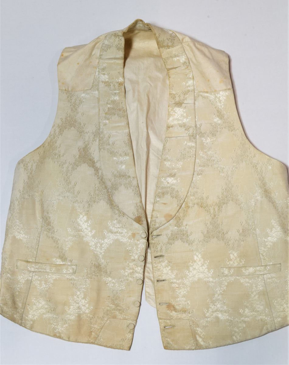 Herrevest i lys beige damask av bomull(?). Sjalskrage med stikning langs kanten. Forstykkene er avrundet nederst og begge har reparasjoner. Lukkes med fire overtrekte knapper. Snittsøm og urlomme på hvert forstykke. Skuldre og rygg er sydd av annet bomullstsoff. I ryggen er det avrundet splitt, spensel med metallspenne. Bomullsfor. På innsiden i ryggen er det små initialer brodert med rød tråd.