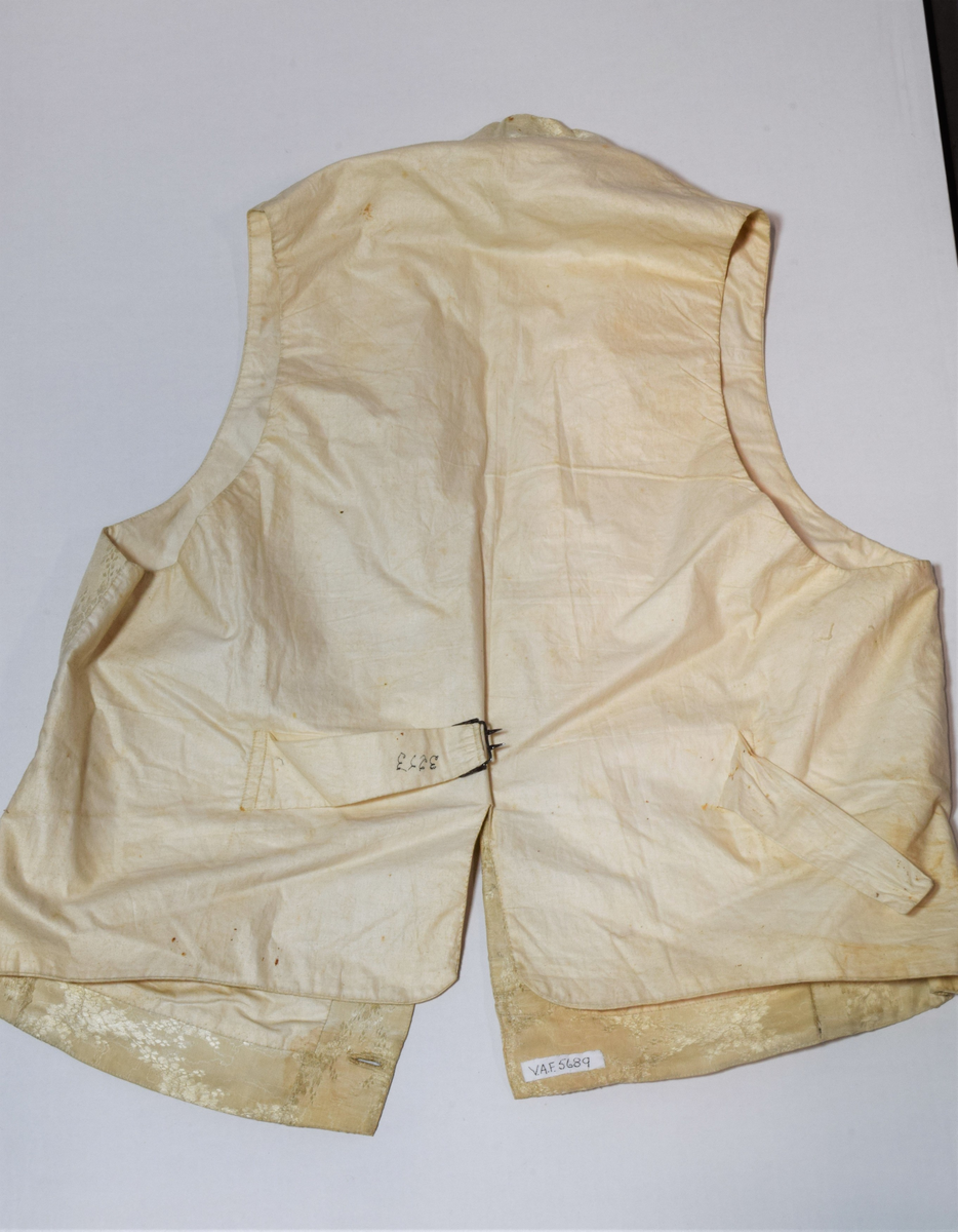 Herrevest i lys beige damask av bomull(?). Sjalskrage med stikning langs kanten. Forstykkene er avrundet nederst og begge har reparasjoner. Lukkes med fire overtrekte knapper. Snittsøm og urlomme på hvert forstykke. Skuldre og rygg er sydd av annet bomullstsoff. I ryggen er det avrundet splitt, spensel med metallspenne. Bomullsfor. På innsiden i ryggen er det små initialer brodert med rød tråd.