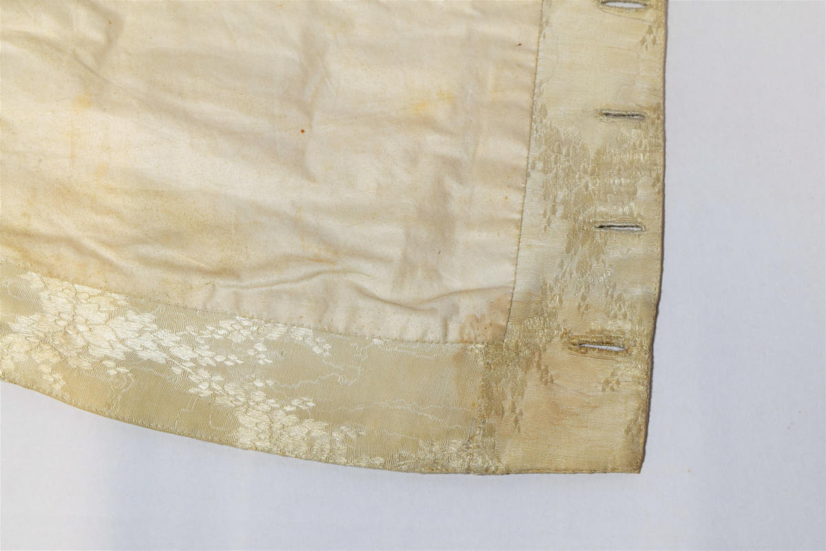 Herrevest i lys beige damask av bomull(?). Sjalskrage med stikning langs kanten. Forstykkene er avrundet nederst og begge har reparasjoner. Lukkes med fire overtrekte knapper. Snittsøm og urlomme på hvert forstykke. Skuldre og rygg er sydd av annet bomullstsoff. I ryggen er det avrundet splitt, spensel med metallspenne. Bomullsfor. På innsiden i ryggen er det små initialer brodert med rød tråd.