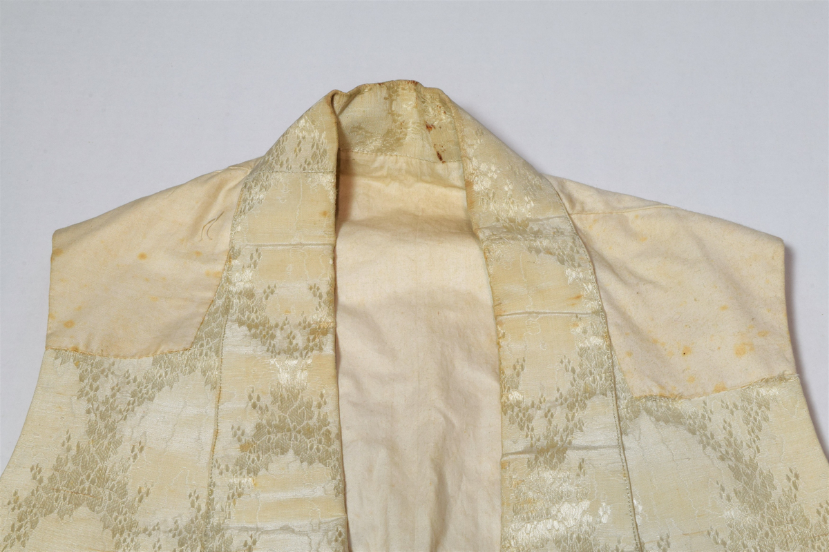 Herrevest i lys beige damask av bomull(?). Sjalskrage med stikning langs kanten. Forstykkene er avrundet nederst og begge har reparasjoner. Lukkes med fire overtrekte knapper. Snittsøm og urlomme på hvert forstykke. Skuldre og rygg er sydd av annet bomullstsoff. I ryggen er det avrundet splitt, spensel med metallspenne. Bomullsfor. På innsiden i ryggen er det små initialer brodert med rød tråd.