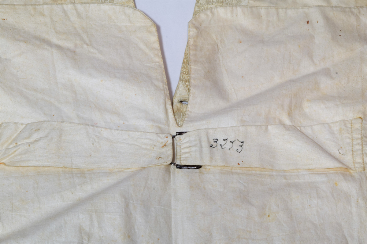 Herrevest i lys beige damask av bomull(?). Sjalskrage med stikning langs kanten. Forstykkene er avrundet nederst og begge har reparasjoner. Lukkes med fire overtrekte knapper. Snittsøm og urlomme på hvert forstykke. Skuldre og rygg er sydd av annet bomullstsoff. I ryggen er det avrundet splitt, spensel med metallspenne. Bomullsfor. På innsiden i ryggen er det små initialer brodert med rød tråd.