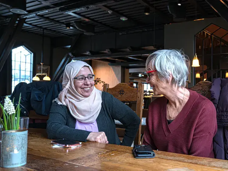 To damer sitter ved et bord i samtale med hverandre. Begge har lagt sine mobiler på bordet. Hun til venstre bærer hijab, hun til høyre har røde briller og grått hår. Et vindu lyser i bakgrunnen