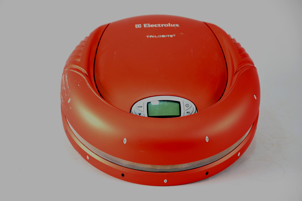 Robotdammsugare typ Trilobite med hölje i röd plast. Dammsugaren navigerar med hjälp av ultraljud. Den återvänder till laddstationen för laddning och stänger av sig själv när dammsugningen är klar.
Märkt "Sample only - not for approval". Typ TR1 ser nr 34700118 model nr ZA1(PS2).