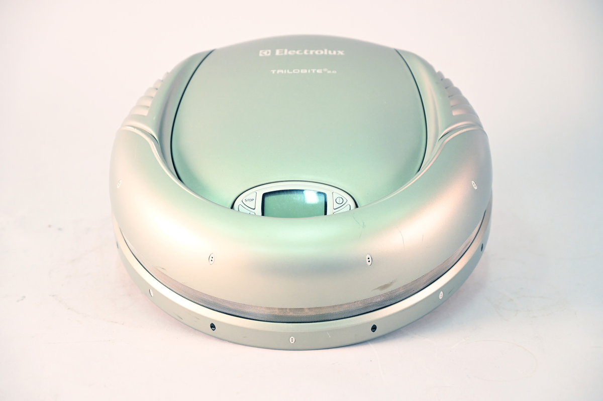 Robotdammsugare typ Trilobite med hölje i grå plast. Dammsugaren navigerar med hjälp av ultraljud. Den återvänder till laddstationen för laddning och stänger av sig själv när dammsugningen är klar.
Typ TR1 ser nr 41700066 model nr ZA2 prod nr 907 350 502.