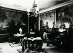 Interiören av Hantverkshuset år 1908. Det finns bord, piano, soffa och väggmålningar.