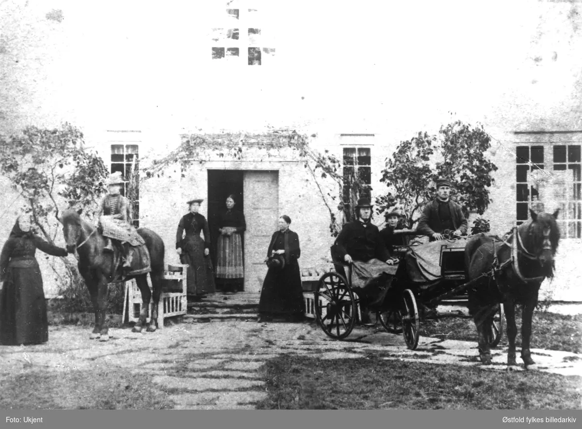 Sokneprest Ole Ulsten med flere ca. 1898-1900. Ulsteen var sokneprest i Tune fra 1898 til 1911. Soknepresten sitter i trilla, ved hans side datteren Inga Ulsten. På hesteryggen til venstre datter Signe Ulsten. Prestefruen, Betsy Marie, til høyre for døren. Ellers folk på gården. 
Det er oppgitt at bildet er fra Tune prestegård, det kan ikke stemme.