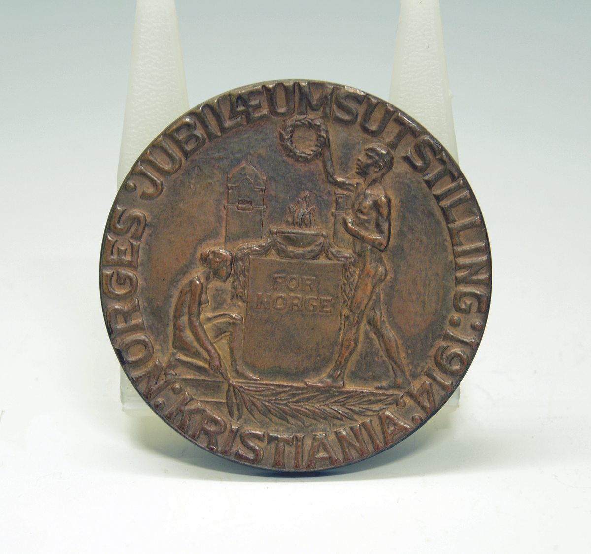 Bronsemedalje fra Jubileumsutstillingen 1914 i eske. Advers: I senter en ildskål på et podium. På venstre side en knelende kvinne med et palmeblad i sin høyre hånd. På høyre siden en stående mann med krans i sin høyre hånd. I bakgrunnen et tårn. Langs kanten innskriften: "Norges jubilæumsutstilling 1914 :Kristiania:" Revers: I senter det norske riksvåpenet, under et banner med innskrift. På hver side av riksvåpenet furukvister med kongler og over "1814-1914".