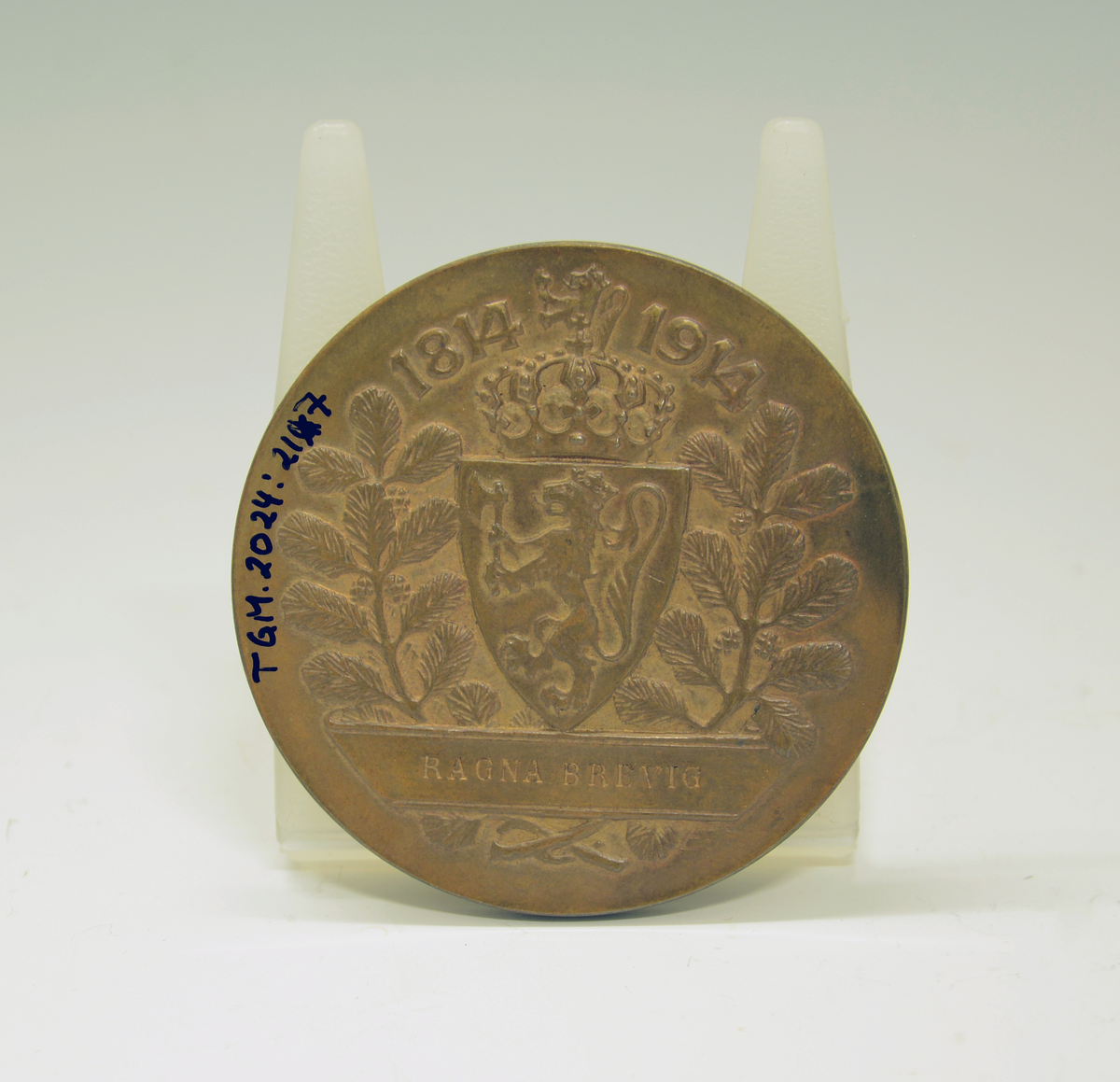 Bronsemedalje fra Jubileumsutstillingen 1914 i eske. Advers: I senter en ildskål på et podium. På venstre side en knelende kvinne med et palmeblad i sin høyre hånd. På høyre siden en stående mann med krans i sin høyre hånd. I bakgrunnen et tårn. Langs kanten innskriften: "Norges jubilæumsutstilling 1914 :Kristiania:" Revers: I senter det norske riksvåpenet, under et banner med innskrift. På hver side av riksvåpenet furukvister med kongler og over "1814-1914".
