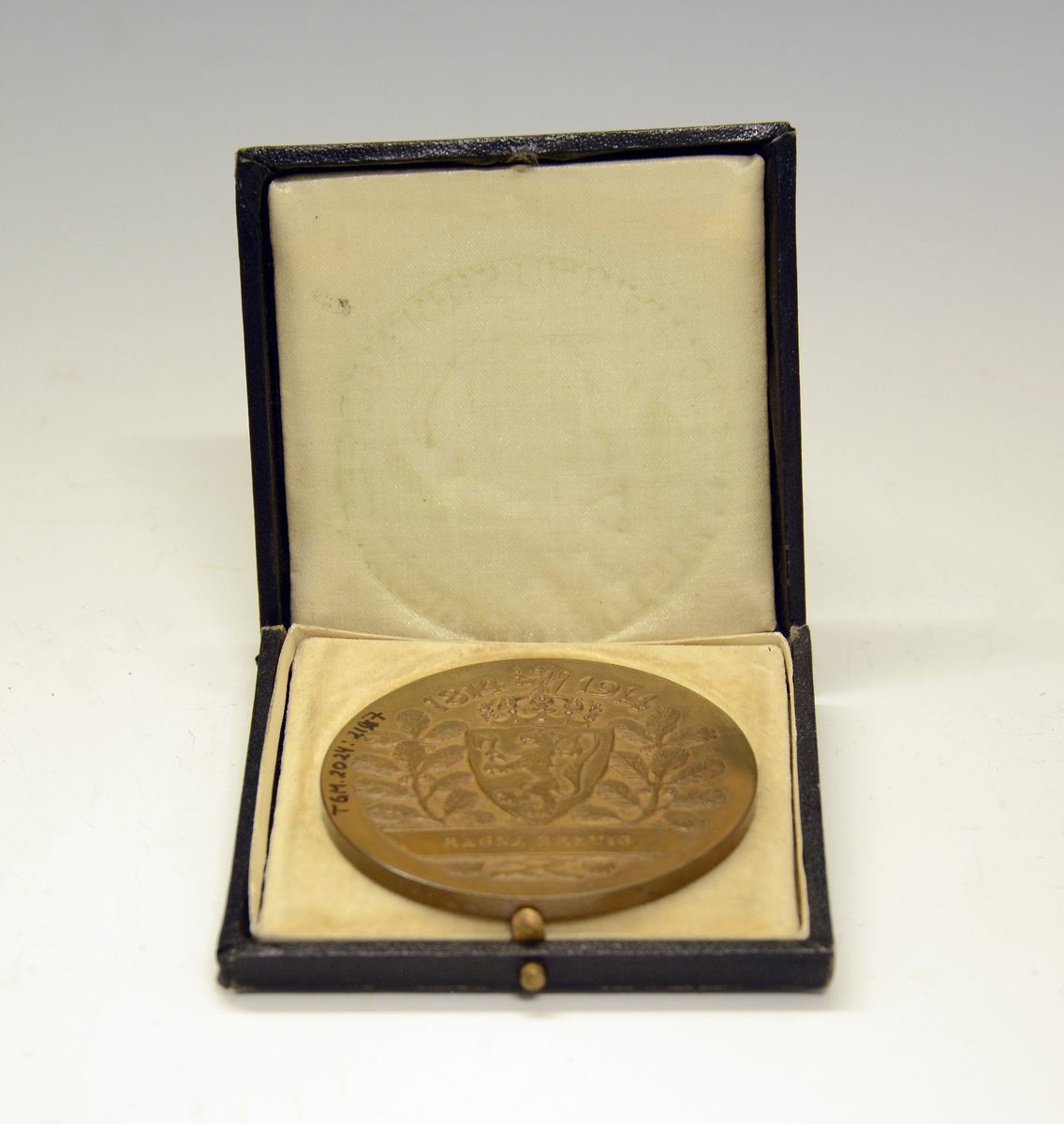 Bronsemedalje fra Jubileumsutstillingen 1914 i eske. Advers: I senter en ildskål på et podium. På venstre side en knelende kvinne med et palmeblad i sin høyre hånd. På høyre siden en stående mann med krans i sin høyre hånd. I bakgrunnen et tårn. Langs kanten innskriften: "Norges jubilæumsutstilling 1914 :Kristiania:" Revers: I senter det norske riksvåpenet, under et banner med innskrift. På hver side av riksvåpenet furukvister med kongler og over "1814-1914".