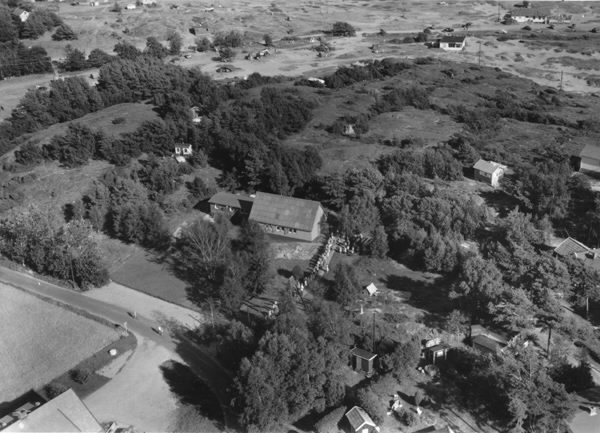 Flygbild som vykort, "S:t Lars kapell, Åsa. Flygfoto." Vy över kapellet och människor på rad utanför. Arkitekt var Johannes Olivegren som vunnit en småkyrkotävling 1954 och kapellet uppfördes i juni 1955. S:t Lars kyrka (som den benämns idag) blev den första av totalt arton kyrkor som Olivegren skulle komma göra ritningarna till. I början av 1950-talet byggdes flera småkyrkor i Göteborg av överblivna militärbaracker och så även S:t Lars. Stommen består av en barack som tidigare tjänstgjort som manskapsbod på Hisingen och i övrigt av en enkel träkonstruktion i gran.
