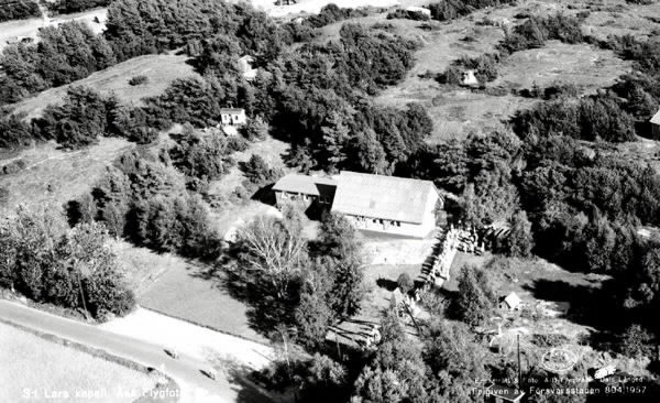 Flygbild som vykort, "S:t Lars kapell, Åsa. Flygfoto." Vy över kapellet och människor på rad utanför. Arkitekt var Johannes Olivegren som vunnit en småkyrkotävling 1954 och kapellet uppfördes i juni 1955. S:t Lars kyrka (som den benämns idag) blev den första av totalt arton kyrkor som Olivegren skulle komma göra ritningarna till. I början av 1950-talet byggdes flera småkyrkor i Göteborg av överblivna militärbaracker och så även S:t Lars. Stommen består av en barack som tidigare tjänstgjort som manskapsbod på Hisingen och i övrigt av en enkel träkonstruktion i gran.