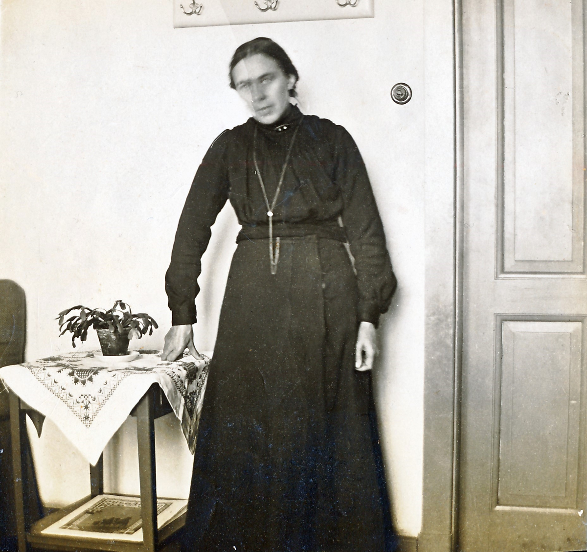 "En för...d kvinna." (Delvis oläslig text.) En mörkt klädd kvinna med lång halsdedja står vid ett bord, täckt av en duk med hallandssöm. På bordet står en novemberkaktus och över kvinnan sitter tre dubbelkrokar.