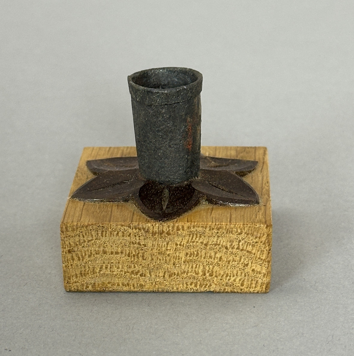 Kvadratisk ljusstake av ek, dekorerad med en snidad värmlandsblomma av mörkt träslag (liknar valnöt) och på denna en smidd ljuspipa med vikt kant.