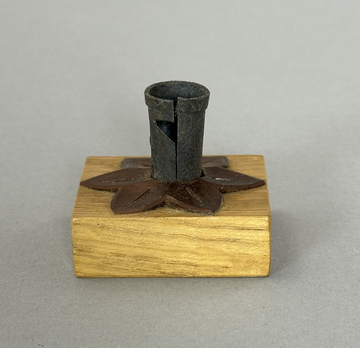 Kvadratisk ljusstake av ek, dekorerad med en snidad värmlandsblomma av mörkt träslag (liknar valnöt) och på denna en smidd ljuspipa med vikt kant.