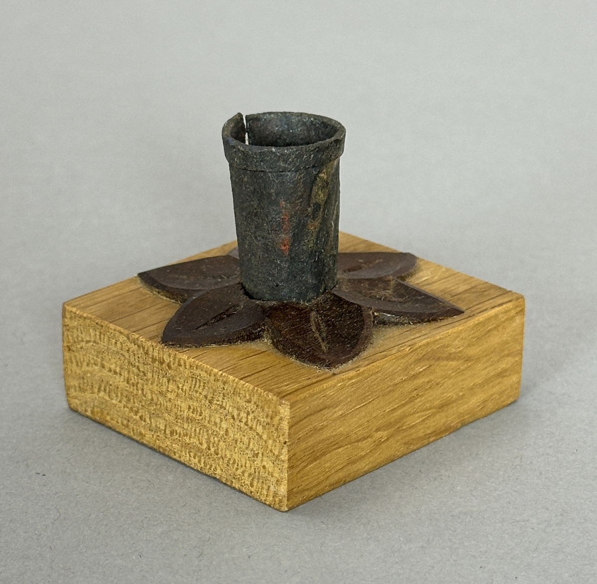 Kvadratisk ljusstake av ek, dekorerad med en snidad värmlandsblomma av mörkt träslag (liknar valnöt) och på denna en smidd ljuspipa med vikt kant.