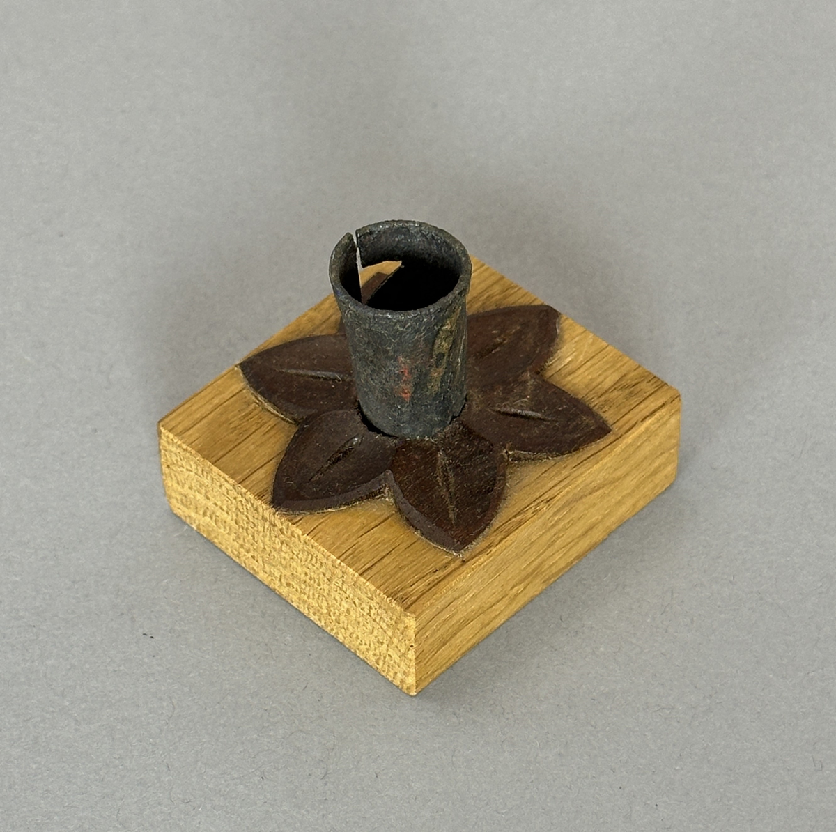 Kvadratisk ljusstake av ek, dekorerad med en snidad värmlandsblomma av mörkt träslag (liknar valnöt) och på denna en smidd ljuspipa med vikt kant.
