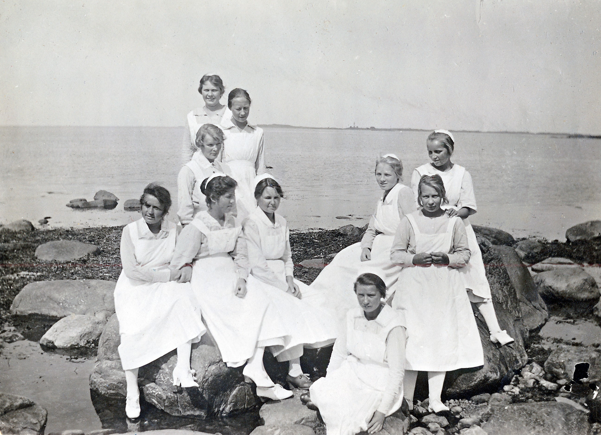 "I kamratkretsen Midsommardagen 1921". Tio unga kvinnor ur personalen vid Kustsanatoriet Apelviken sitter på stenar vid stranden i Lilla Apelviken.